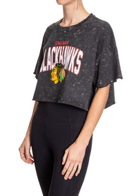 Load image into Gallery viewer, Woman standing sideways in front of a white background wearing an oversized, black, acid wash crop top - featuring a Chicago Blackhawks logo in the center of the shirt.

