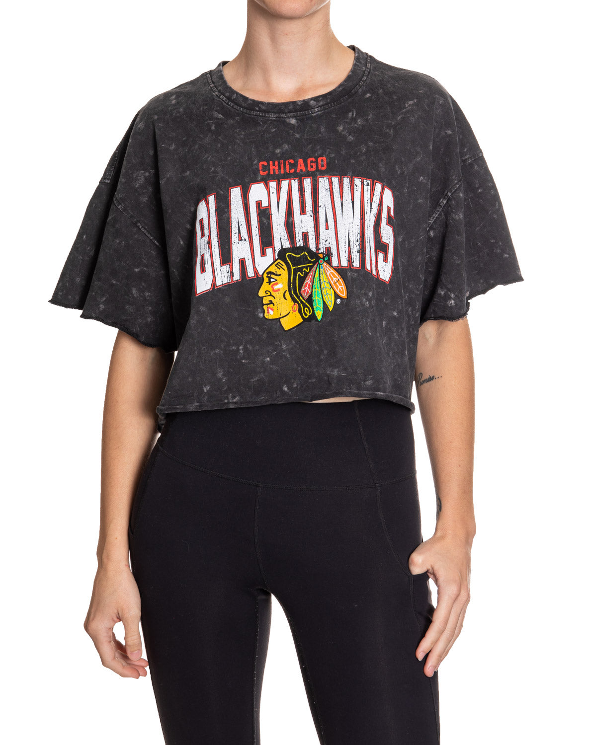 Woman standing in front of a white background, wearing an oversized black acid wash crop top - featuring a Chicago Blackhawks logo in the center of the shirt.