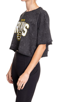 Load image into Gallery viewer, Woman standing sideways in front of a white background wearing an oversized, black, acid wash crop top - featuring a Boston Bruins logo in the center of the shirt.
