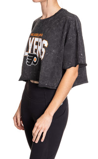 Load image into Gallery viewer, Woman standing sideways in front of a white background wearing an oversized, black, acid wash crop top - featuring a Philadelphia Flyers logo in the center of the shirt.
