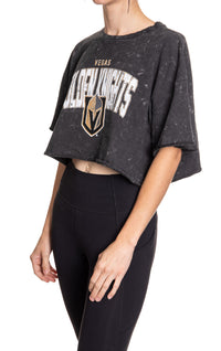 Load image into Gallery viewer, Woman standing sideways in front of a white background wearing an oversized, black, acid wash crop top - featuring a Vegas Golden Knights logo in the center of the shirt.
