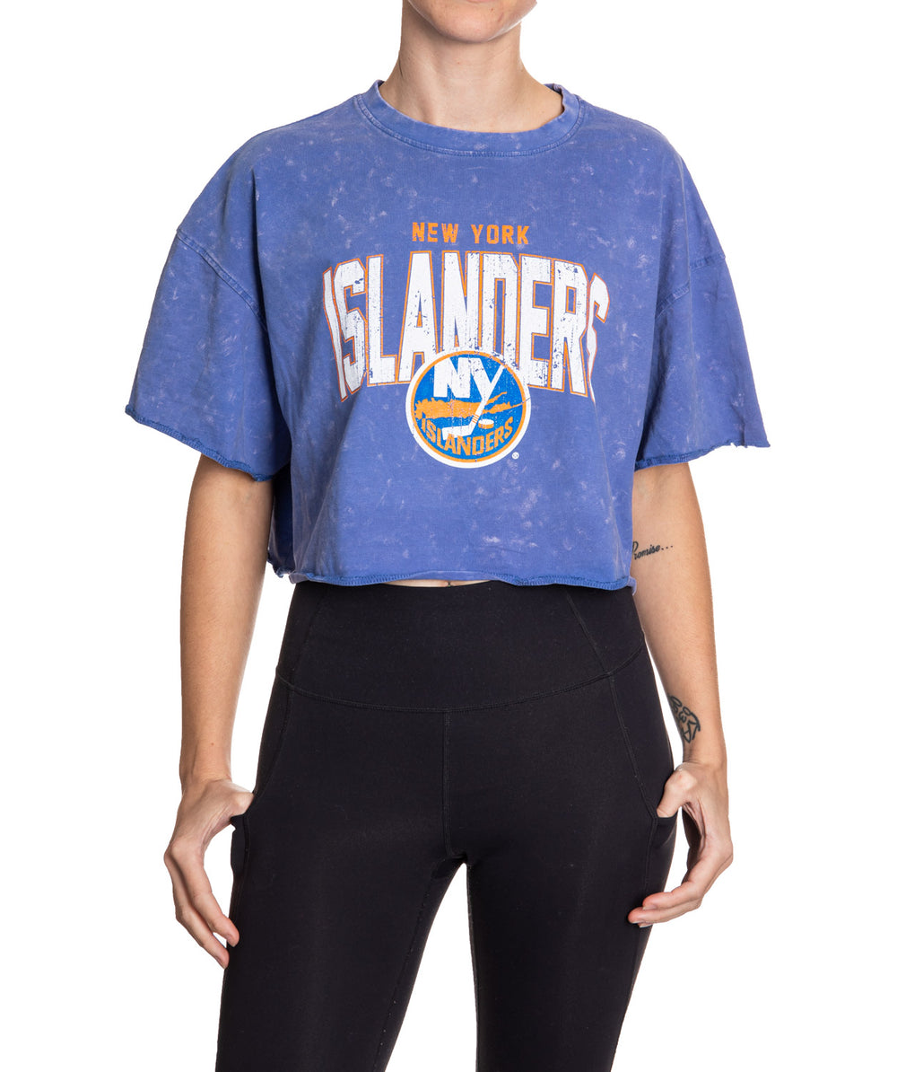 Woman standing in front of a white background wearing an oversized, blue, acid wash crop top - featuring a New York Islanders logo in the center of the shirt.