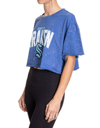 Load image into Gallery viewer, Woman standing sideways in front of a white background wearing an oversized, blue, acid wash crop top - featuring a Seattle Kraken logo in the center of the shirt.
