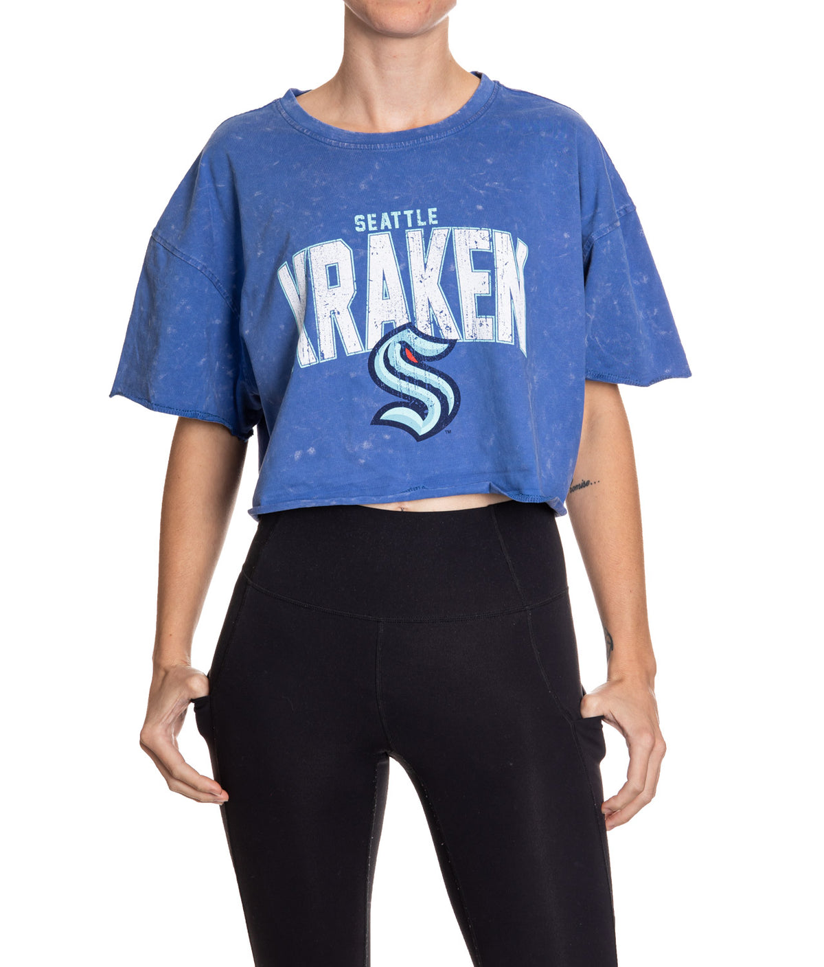 Woman standing in front of a white background wearing an oversized, blue, acid wash crop top - featuring a Seattle Kraken logo in the center of the shirt.
