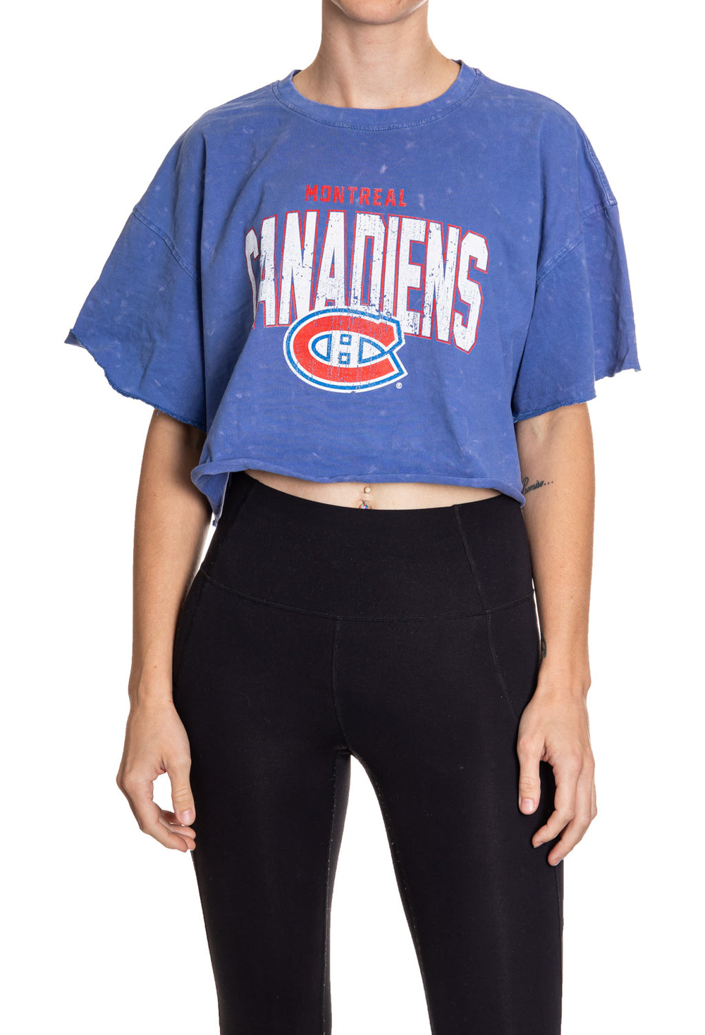 Woman standing in front of a white background wearing an oversized, blue, acid wash crop top - featuring a Montreal Canadians logo in the center of the shirt.