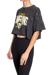 Load image into Gallery viewer, Woman standing sideways in front of a white background wearing an oversized, black, acid wash crop top - featuring a Pittsburgh Penguins logo in the center of the shirt.
