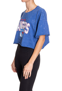 Load image into Gallery viewer, Woman standing sideways in front of a white background wearing an oversized, blue, acid wash crop top - featuring a New York Rangers logo in the center of the shirt.
