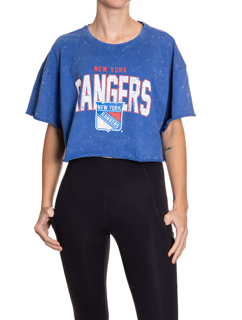 Woman standing in front of a white background wearing an oversized, blue, acid wash crop top - featuring a New York Rangers logo in the center of the shirt.