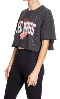 Load image into Gallery viewer, Woman standing sideways in front of a white background wearing an oversized, black, acid wash crop top - featuring a Detroit Red Wings logo in the center of the shirt.
