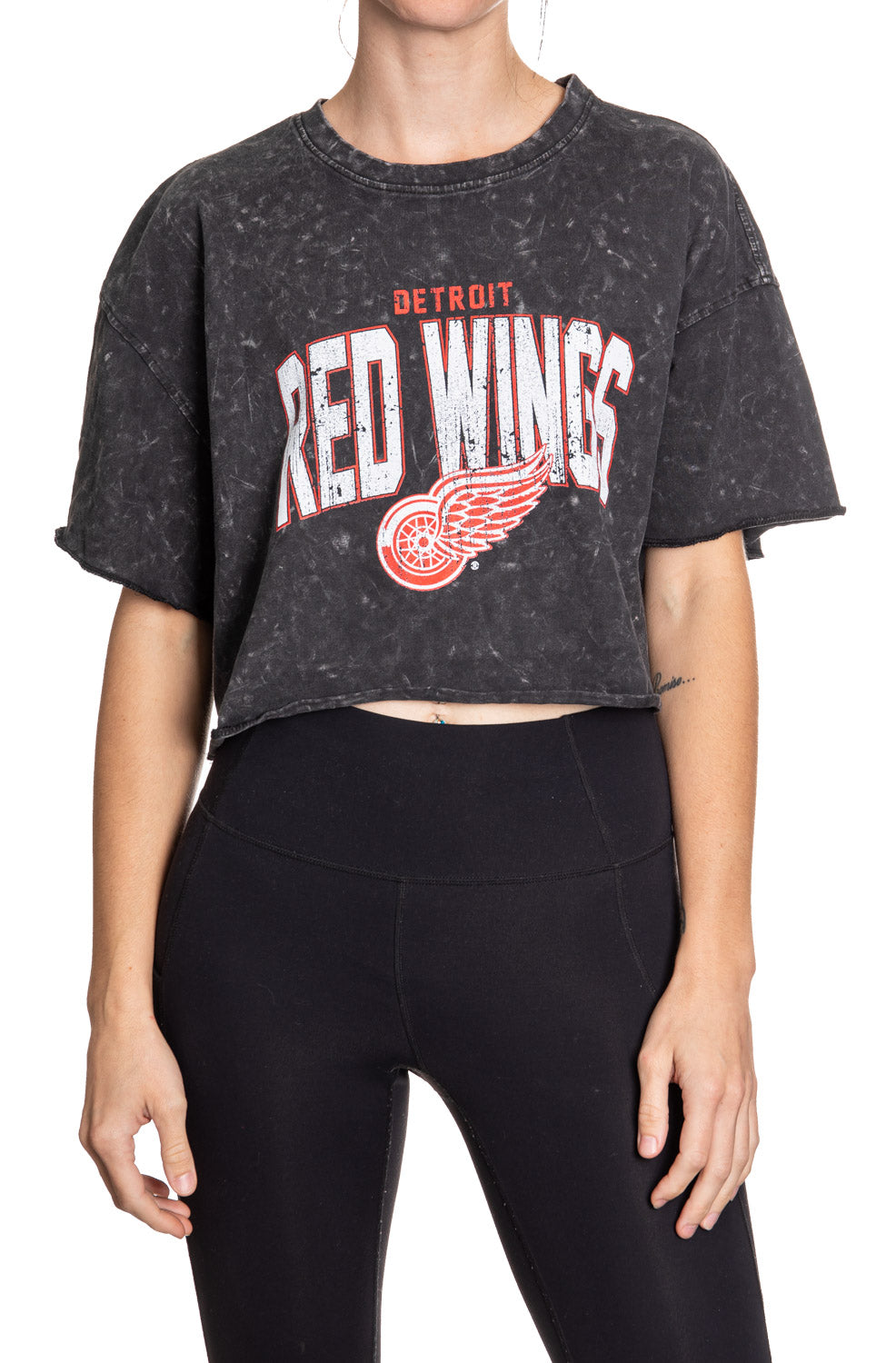 Woman standing in front of a white background wearing an oversized, black, acid wash crop top - featuring a Detroit Red Wings logo in the center of the shirt.