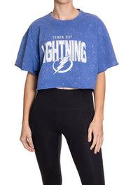 Load image into Gallery viewer, Woman standing in front of a white background wearing an oversized, blue, acid wash crop top - featuring a Tampa Bay Lightning logo in the center of the shirt.
